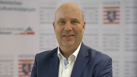 Michael Ashelm lächelt in die Kamera. Er ist bis zur Brust zu sehen, trägt ein blaugraues Sakko und ein weißes Hemd.