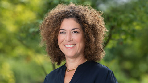 Alice Engel lächelt in die Kamera. Sie trägt eine goldene Kette und eine dunkelblaue Bluse. Im Hintergrund sind grüne Pflanzen zu sehen.