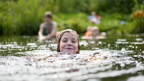 Kind schwimmt glücklich im See. Im Hintergrund kommt auch die Familie ins Wasser.