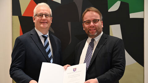 Minister Gremmels (rechts) steht neben Dr. Johannes Kistenich-Zerfaß. Beide halten gemeinsam eine Urkunde in Händen und lächeln in die Kamera.