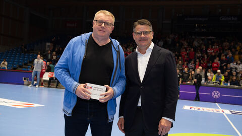 Ministerpräsident Boris Rhein steht auf dem Spielfeld der MT Melsungen. Der Ehrenamtliche Uwe Lohmannhat von ihm eine Auszeichnung überreicht bekommen. Diese hält er in der Hand.