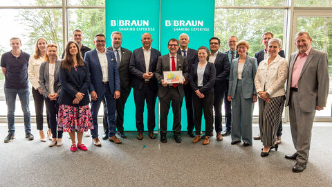 Innenminister Roman Poseck, B. Braun-Vorstandsmitglied Dr. Stefan Ruppert, Nordhessens Polizeipräsident Konrad Stelzenbach und Landtagsabgeordneter Günther Rudolph sowie weitere Mitarbeiterinnen und Mitarbeiter der Firma B. Braun stehen zusammen vor grünem Auftseller