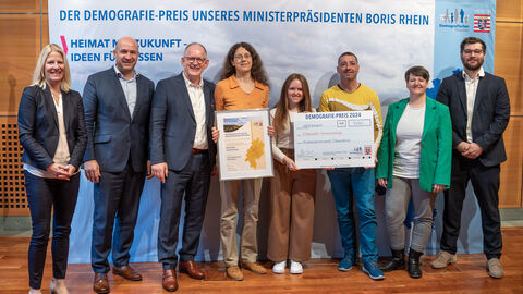 Eine Gruppe Personen steht vor einem Aufsteller mit der Aufschrift "Der Demografie-Preis unseres Ministerpräsidenten Boris Rhein". Eine Person hält eine gerahmte Urkunde in Händen, zwei weitere einen Scheck.