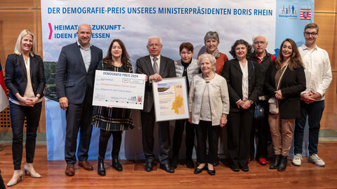 Eine Gruppe Personen steht vor einem Aufsteller mit der Aufschrift "Der Demografie-Preis unseres Ministerpräsidenten Boris Rhein". Eine Person hält eine gerahmte Urkunde in Händen, eine weitere einen Scheck.