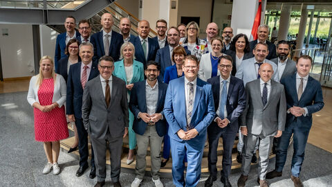 27 Männer und Frauen stehen in Anzügen und Hosenanzügen in einer Eingangshalle.